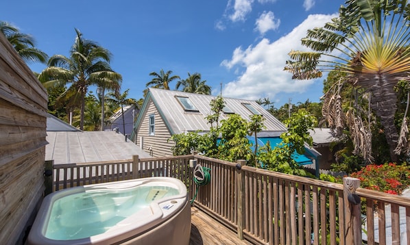 A wooden deck with a hot tub, surrounded by tropical vegetation and a bungalow-style house with a sloped roof.