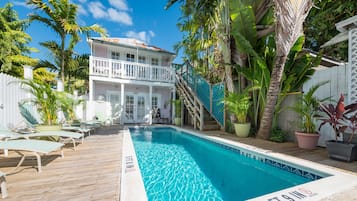 A pool area with a wooden deck, lounge chairs, and a two-story building with a balcony.