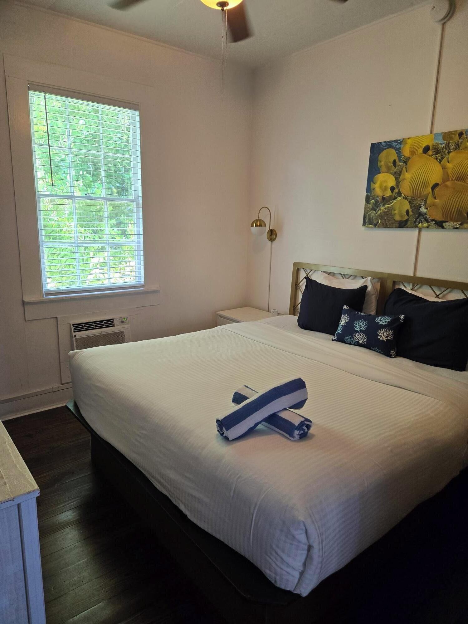 A neatly made bed with a white comforter and two pillows, a bedside lamp, a window with blinds, and a painting on the wall.
