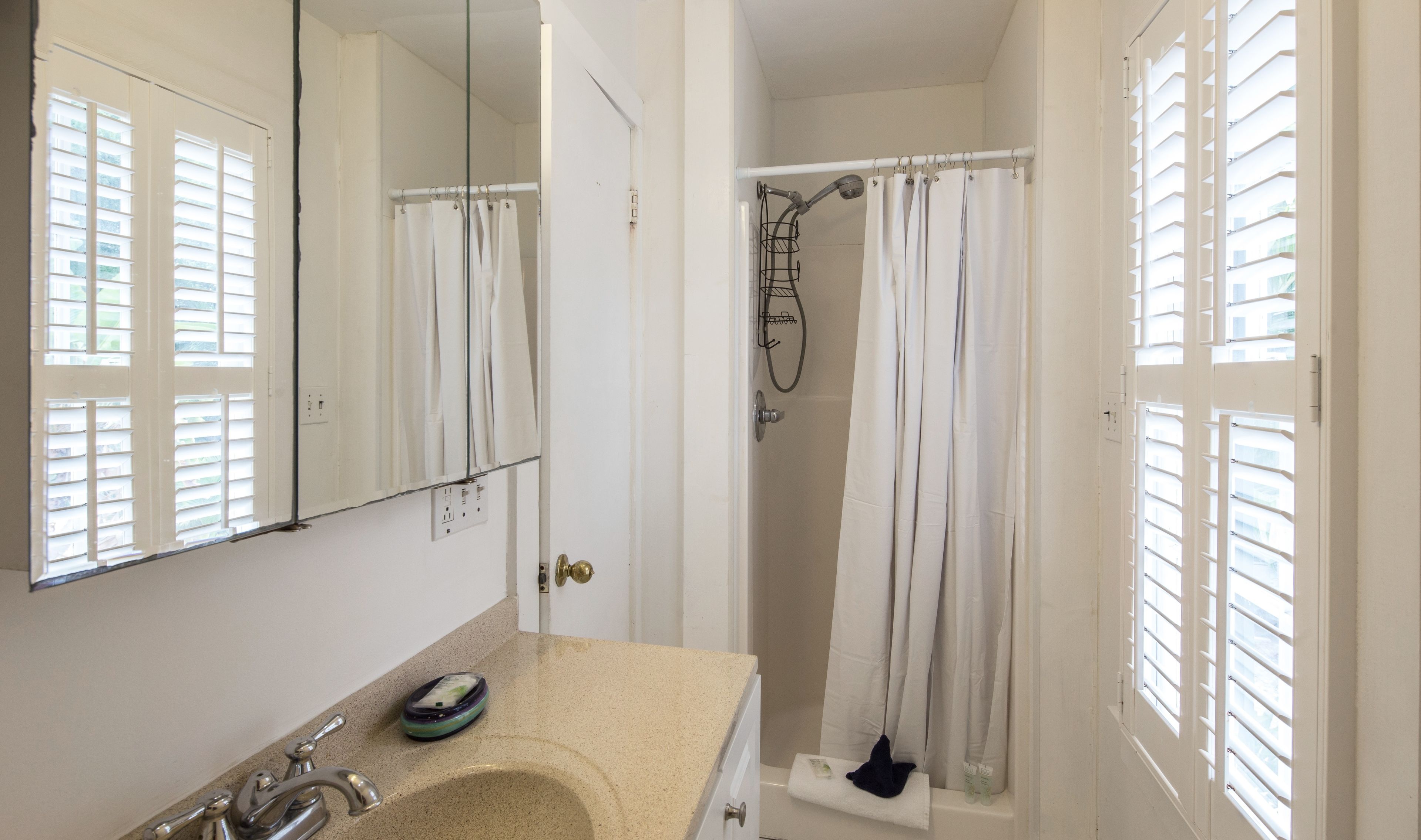 A bathroom with a sink, mirror, shower, and window blinds.