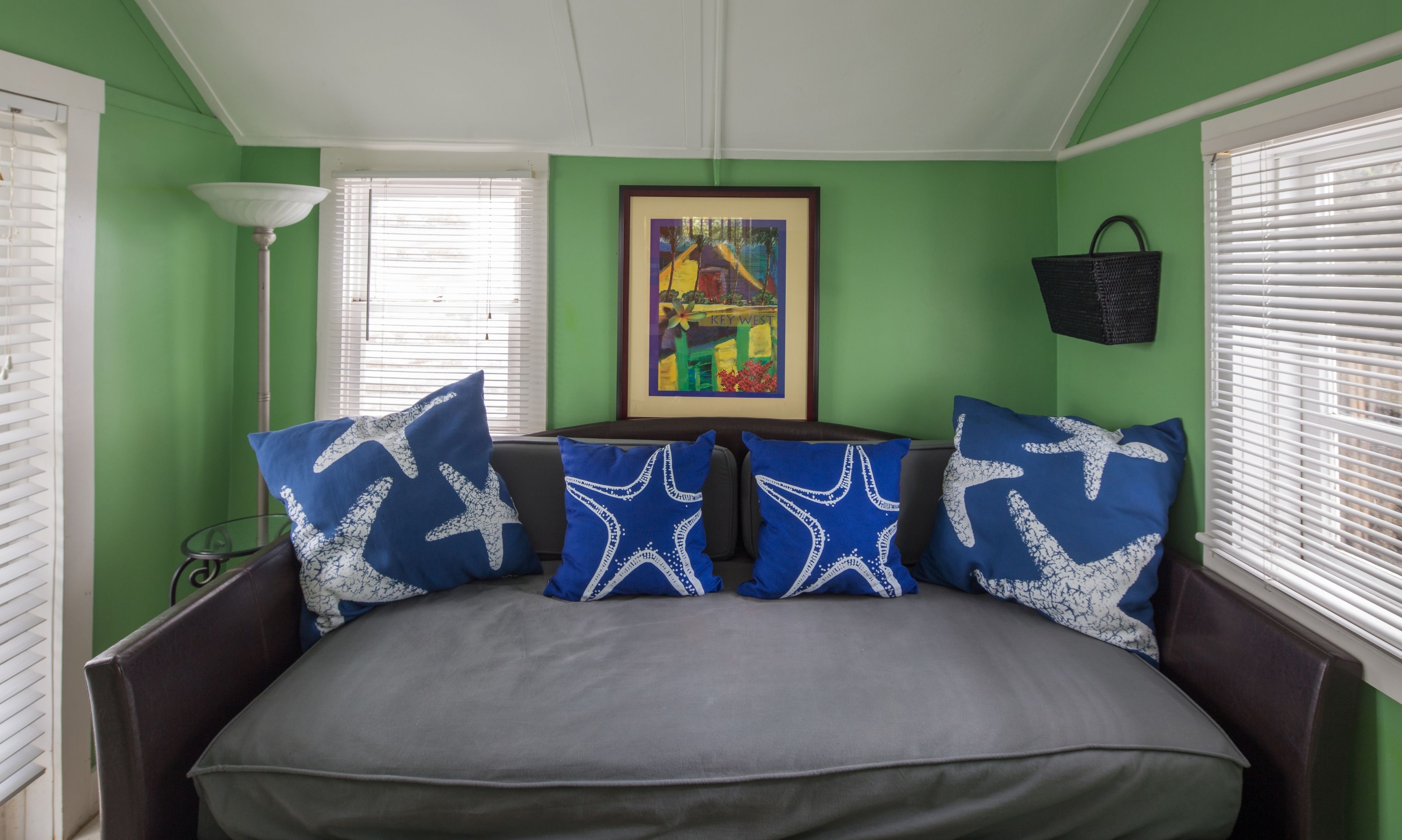 Ein Zimmer mit grünen Wänden, einem Bett mit grauem Topper und blauen Kissen, einem gerahmten Bild an der Wand und weißen Jalousien an den Fenstern.