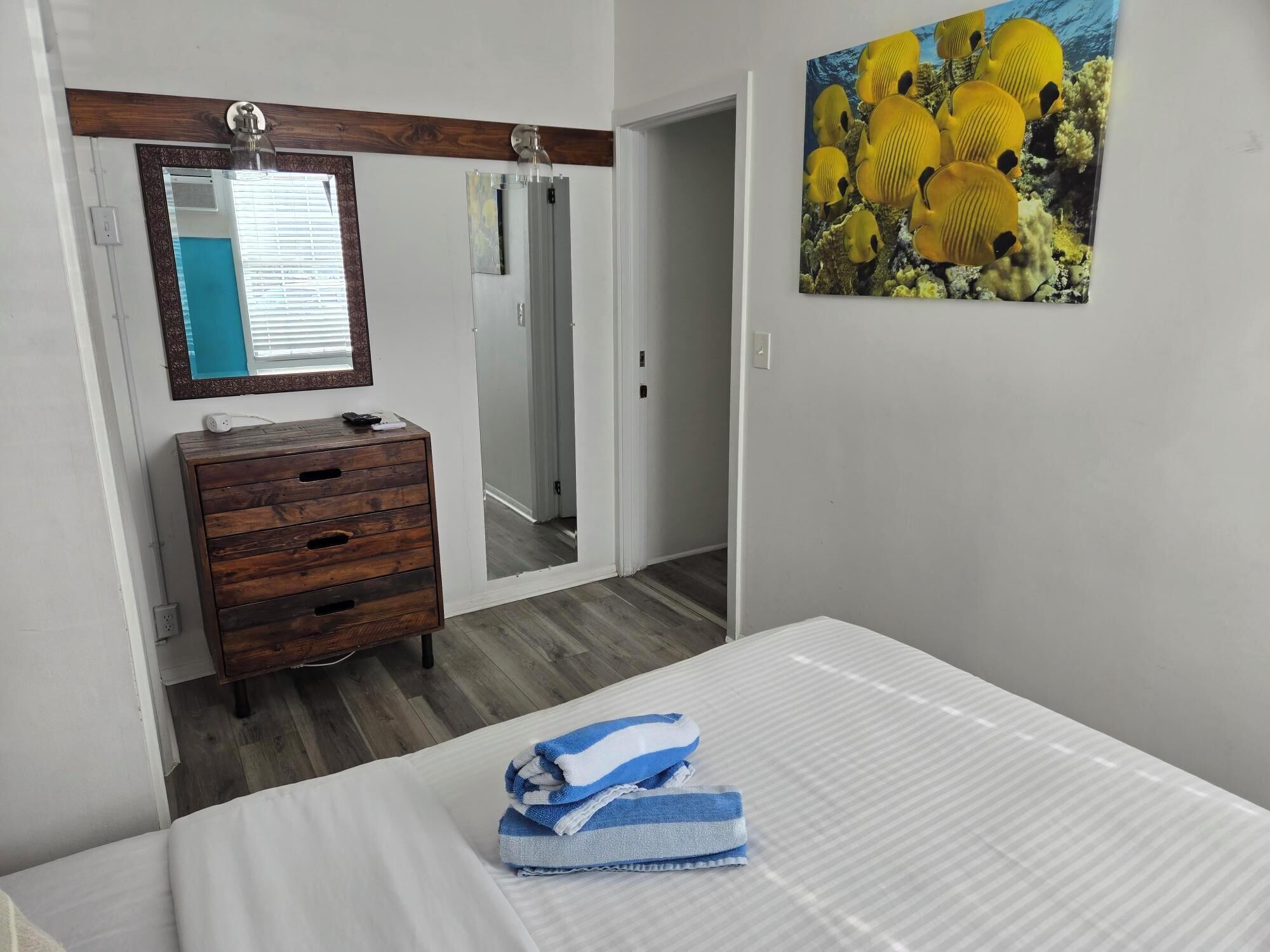 A bedroom with a bed, a wooden dresser, a mirror, and a painting of tropical fish.