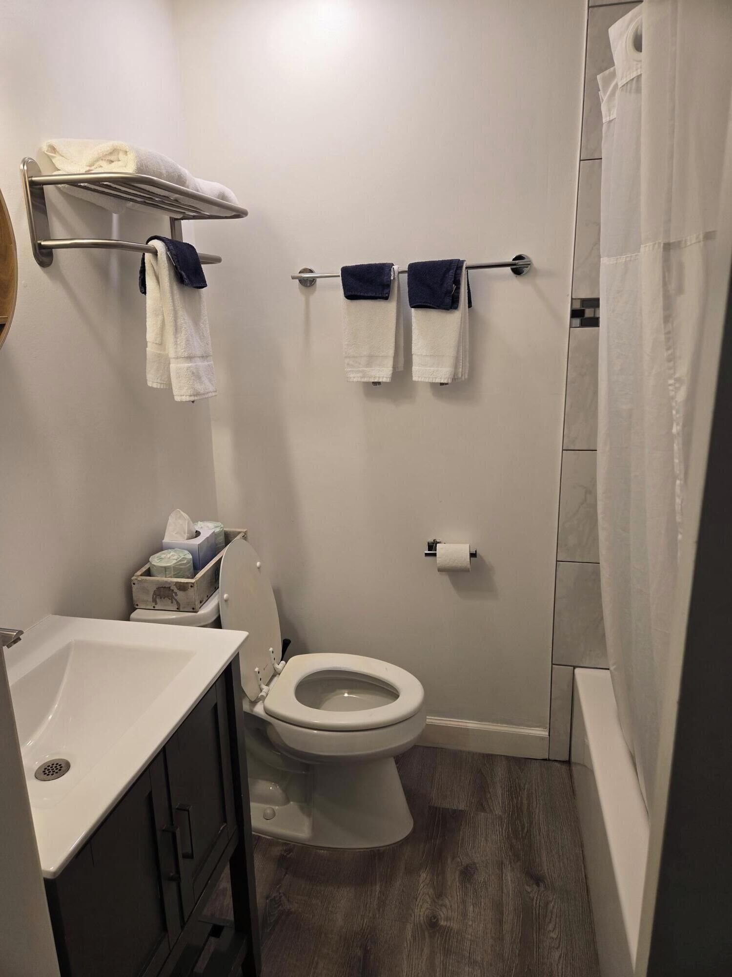 A small bathroom with a toilet, sink, and shower. There are towels hanging on a rack and a shelf above the toilet.