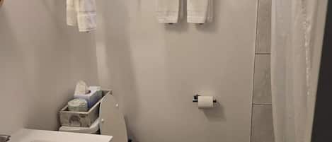 A small bathroom with a toilet, sink, and shower. There are towels hanging on a rack and a shelf above the toilet.