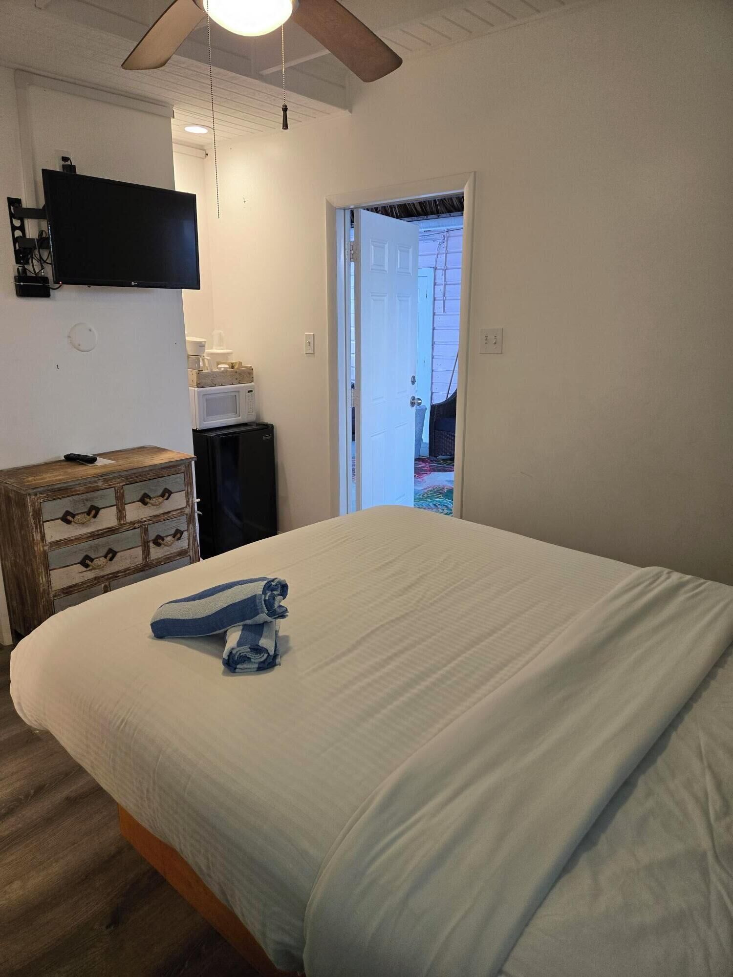 Un dormitorio con una cama, un aparador de madera, un televisor montado en la pared y un ventilador de techo.
