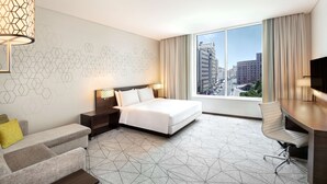 A modern hotel room with a bed, a desk, a chair, and a large window offering a city view.