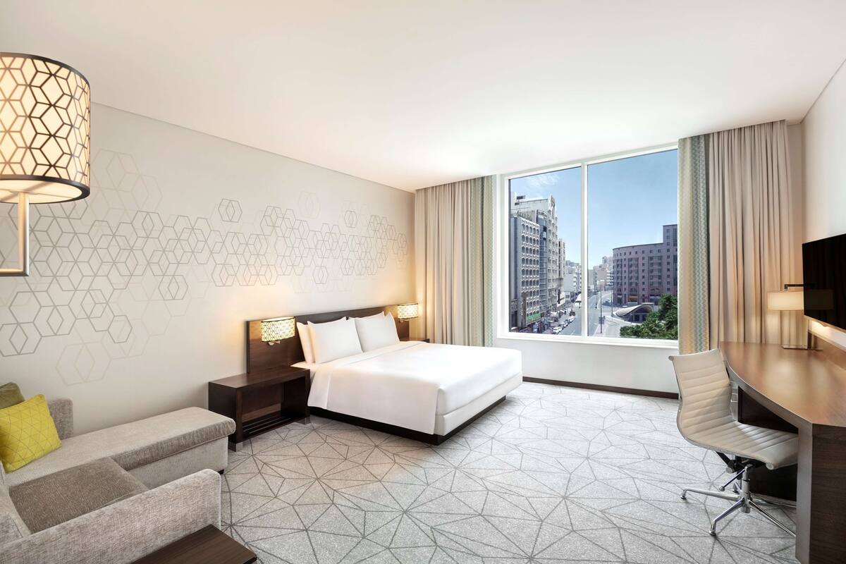 Een moderne hotelkamer met een bed, bureau, stoel en groot raam met uitzicht op de stad.