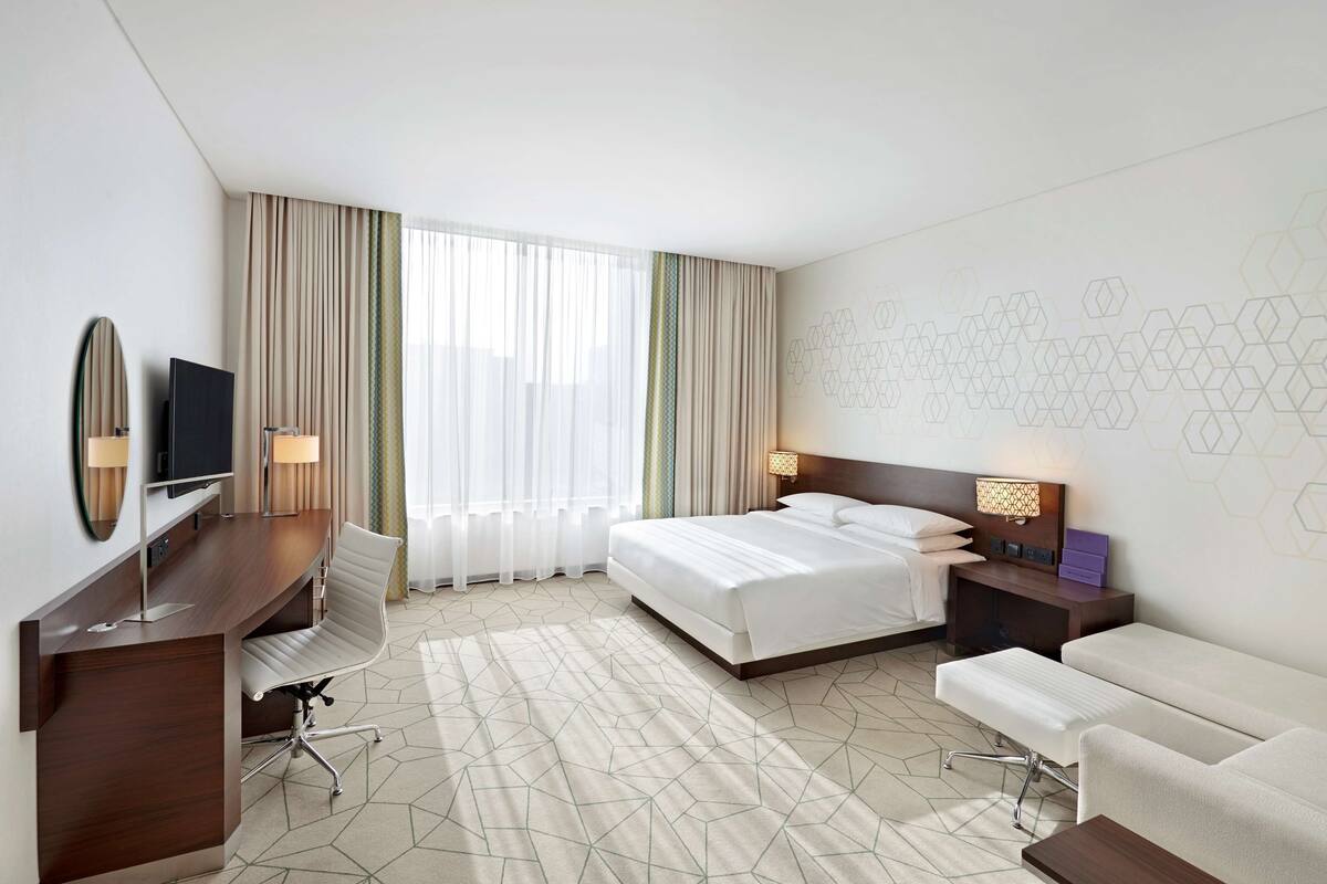 Een hotelkamer met een bed, nachtkastjes, een bureau met stoel, een televisie en een groot raam met gordijnen.