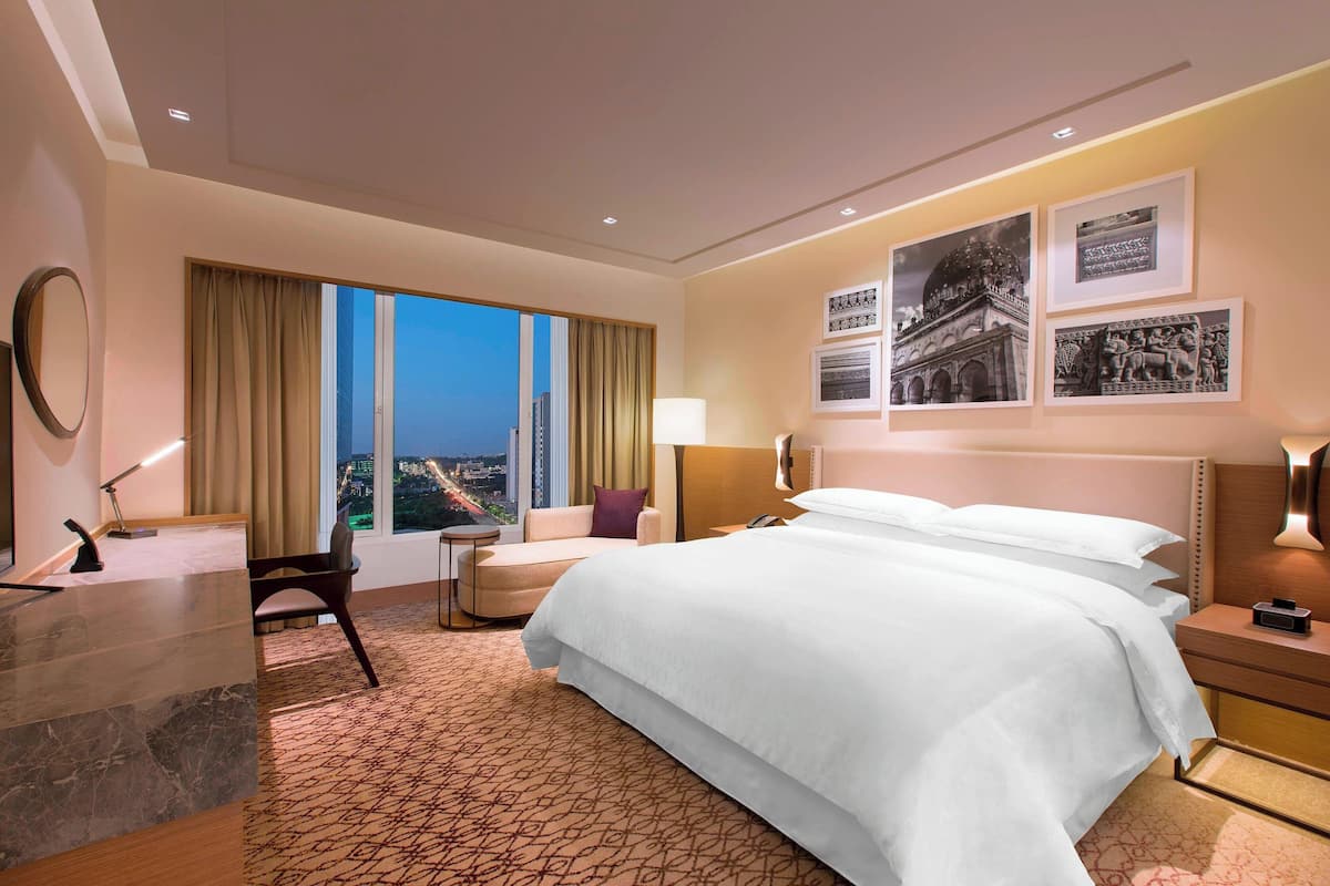 Ein Hotelzimmer mit einem großen Bett, einem Schreibtisch, einem Sessel und Blick auf die Stadt im Abendrot.