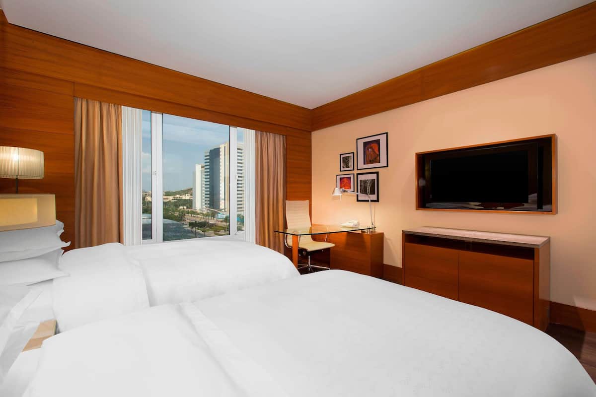 Ein Hotelzimmer mit zwei Betten, einem Schreibtisch mit Stuhl, einem Fernseher und einem Fenster mit Blick auf die Stadt.