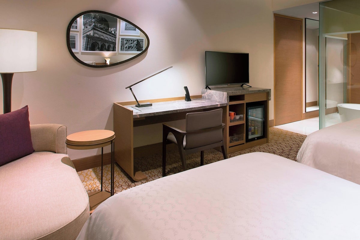 Ein Hotelzimmer mit zwei Betten, einem Schreibtisch mit Computer, einem Stuhl, einem kleinen Tisch, einer Lampe und einem Spiegel.
