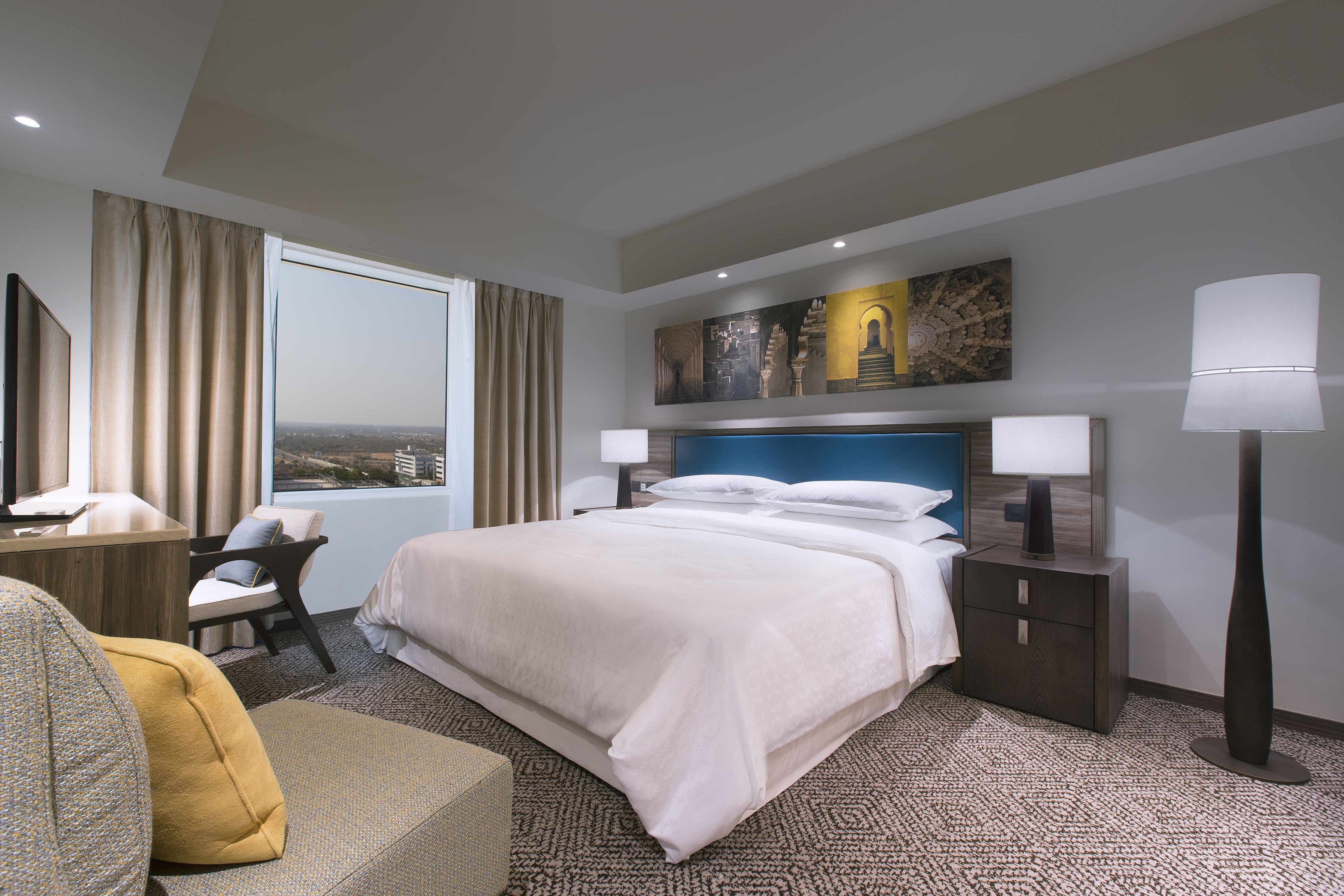 A hotel room with a large bed, a desk, a chair, a lamp, and a view of the city.