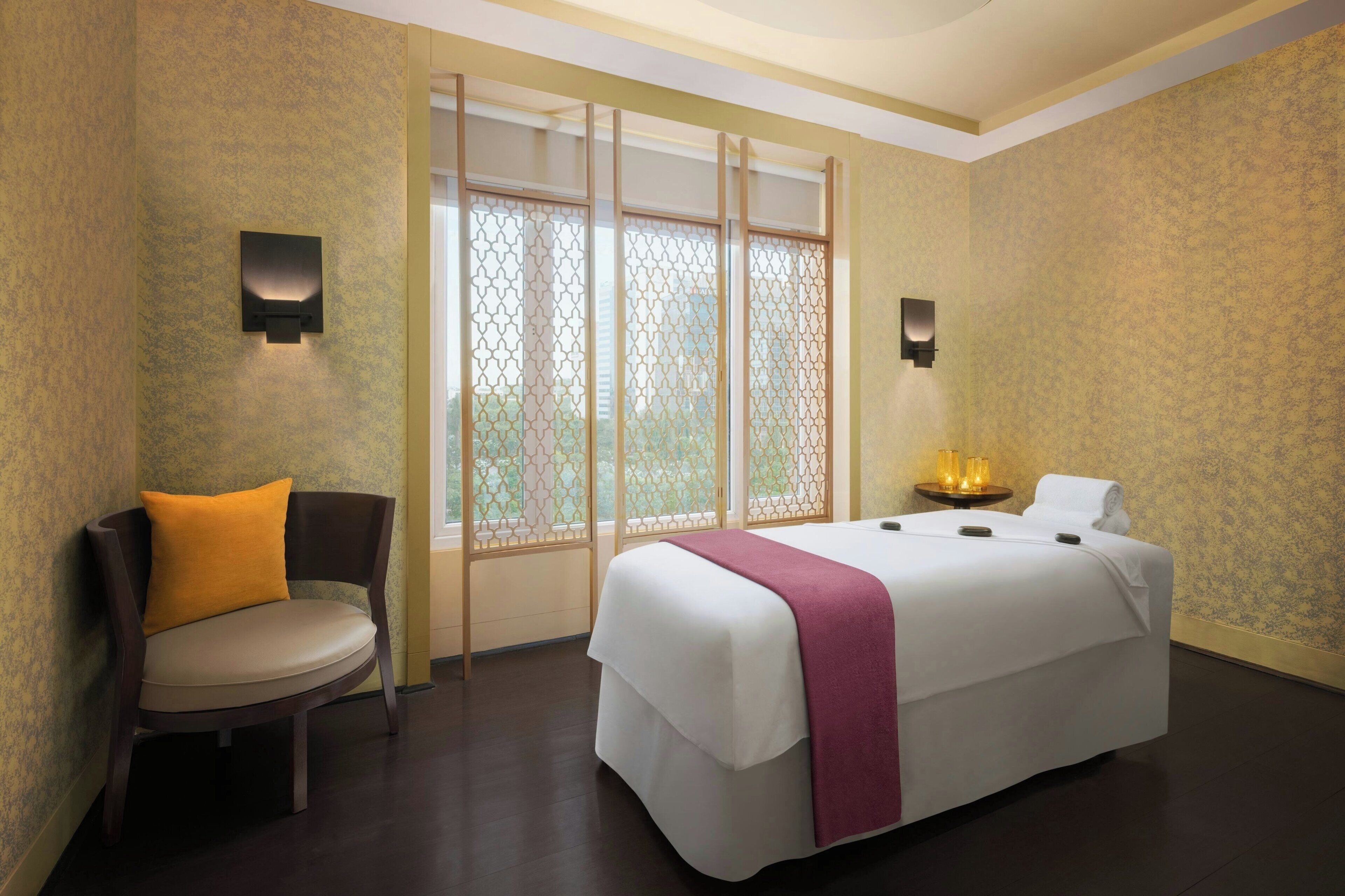 A hotel room with a bed, a chair, a window with a view, and wall-mounted lights.