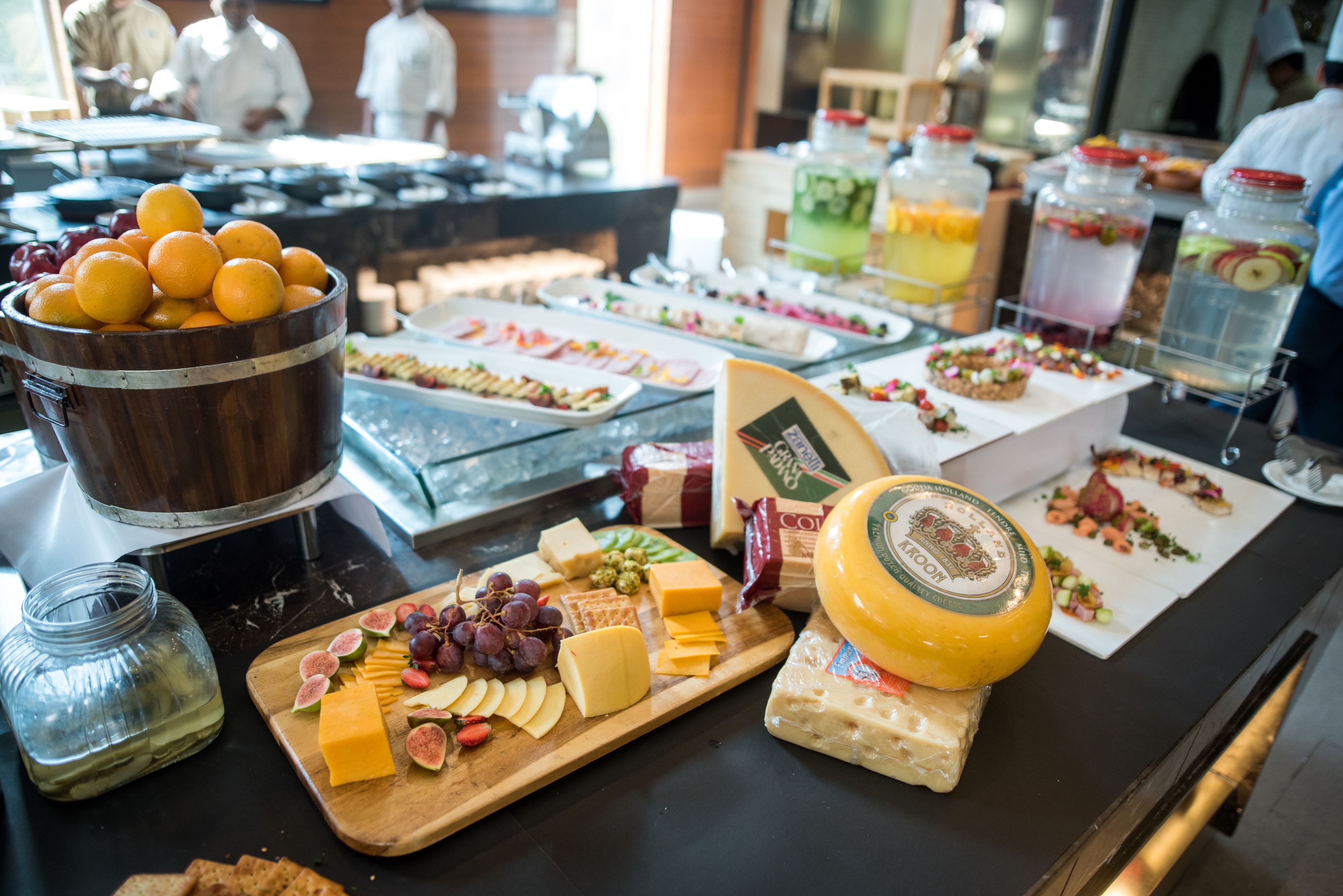 A buffet with various cheeses, fruits, and a wooden bowl of oranges.