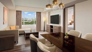 Een moderne hotelkamer met een bank, een eettafel, stoelen, een televisie en een groot raam met uitzicht op de stad.