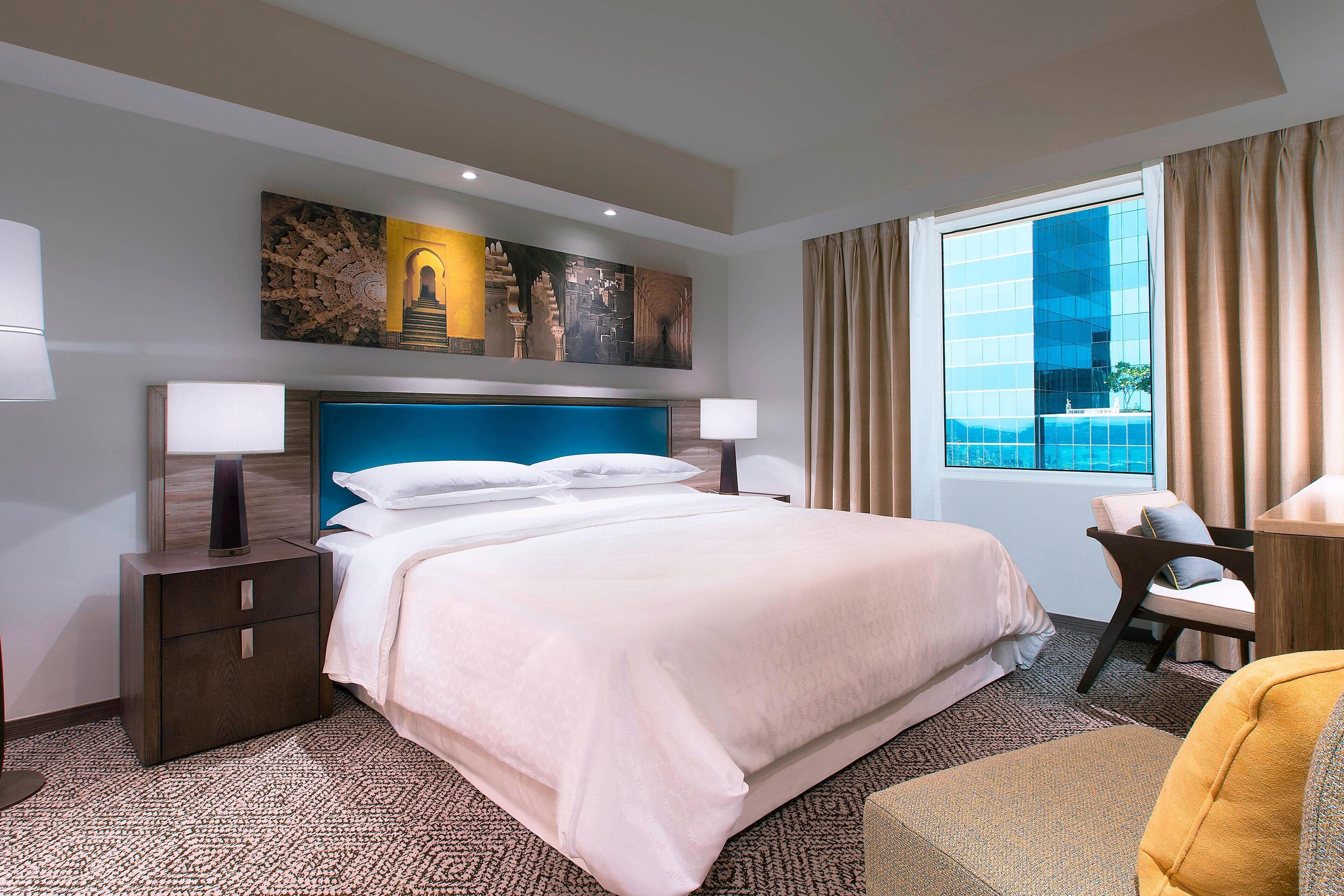 A hotel room with a large bed, two bedside tables with lamps, a chair, and a large window with curtains.