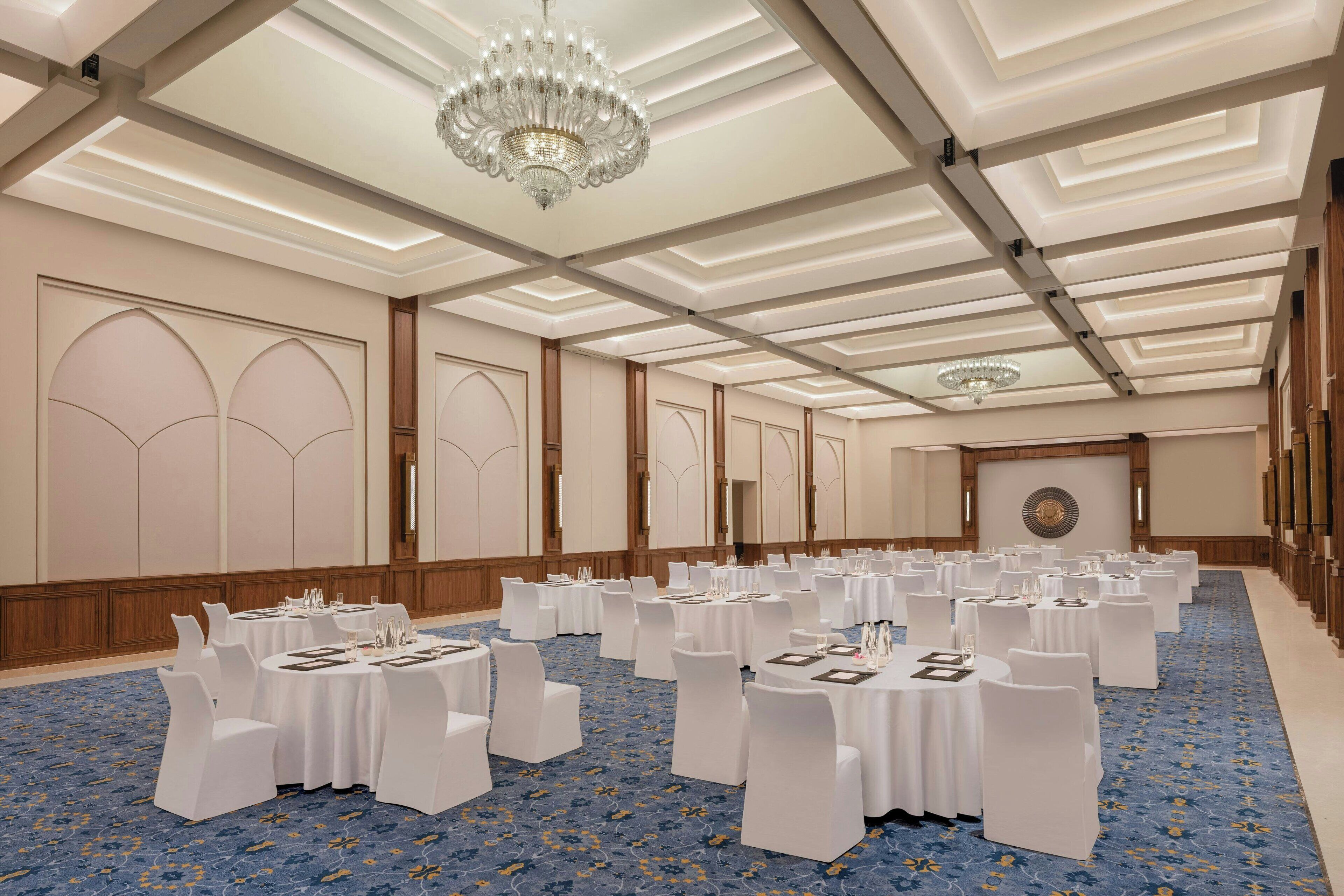 Uma sala de conferências ampla com mesas redondas e cadeiras brancas, um candelabro e um tapete azul.