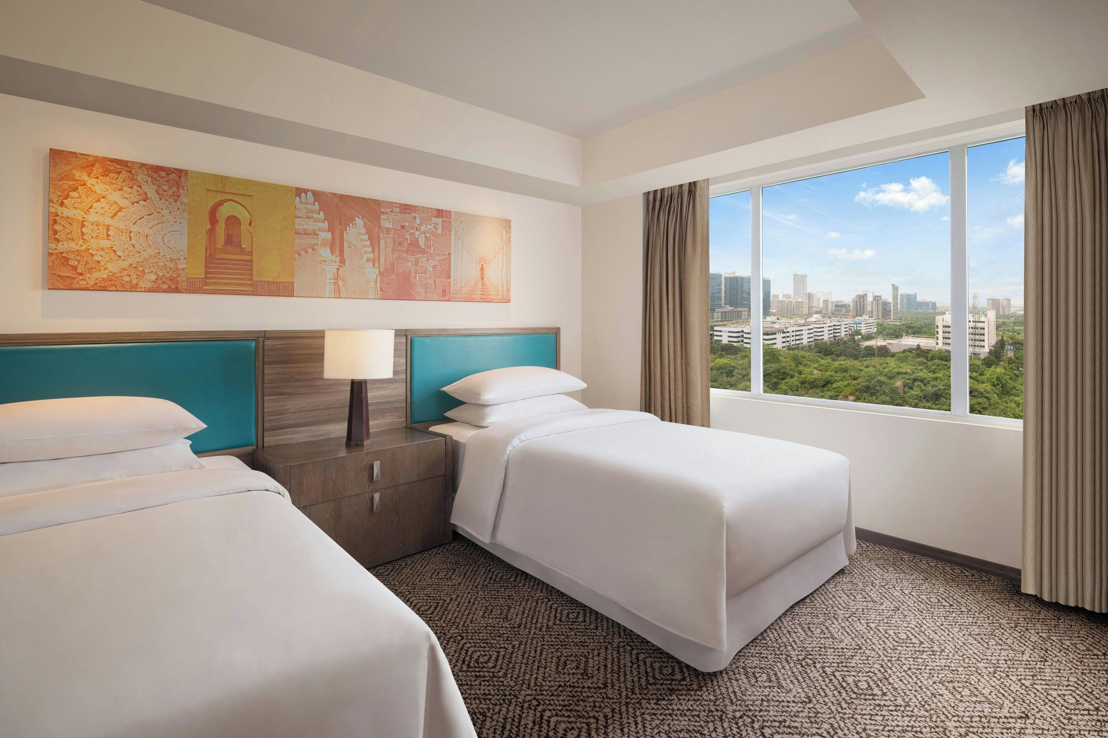 Una habitación de hotel con dos camas, un ventanal con vista a la ciudad, un cabecero con detalles en color turquesa y una obra de arte colgada en la pared.