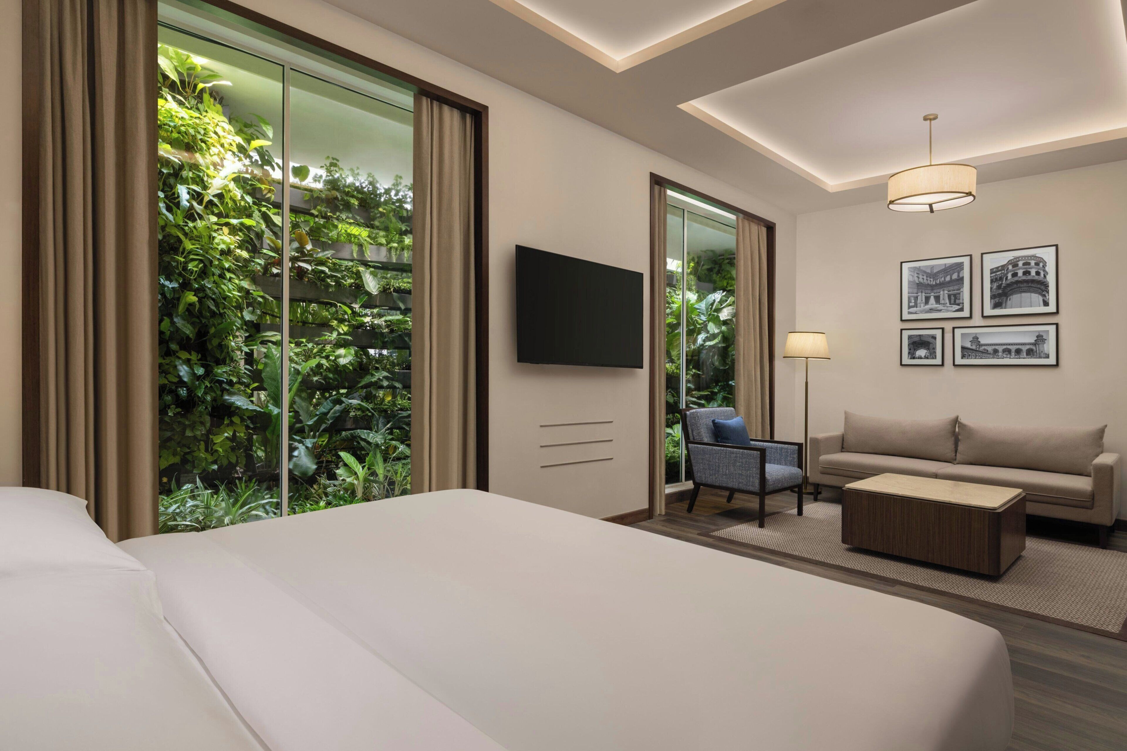 Habitación de hotel con una cama grande, un sofá, una mesa de centro, un televisor y vistas a zonas verdes a través de una puerta corrediza.