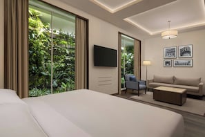 A hotel room with a large bed, a sofa, a coffee table, a TV, and a view of greenery through a sliding door.