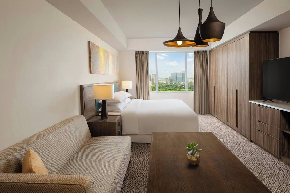 Ein Hotelzimmer mit Bett, einer Sitzecke, einem Fernseher und einem großen Fenster mit Blick auf die Stadt.