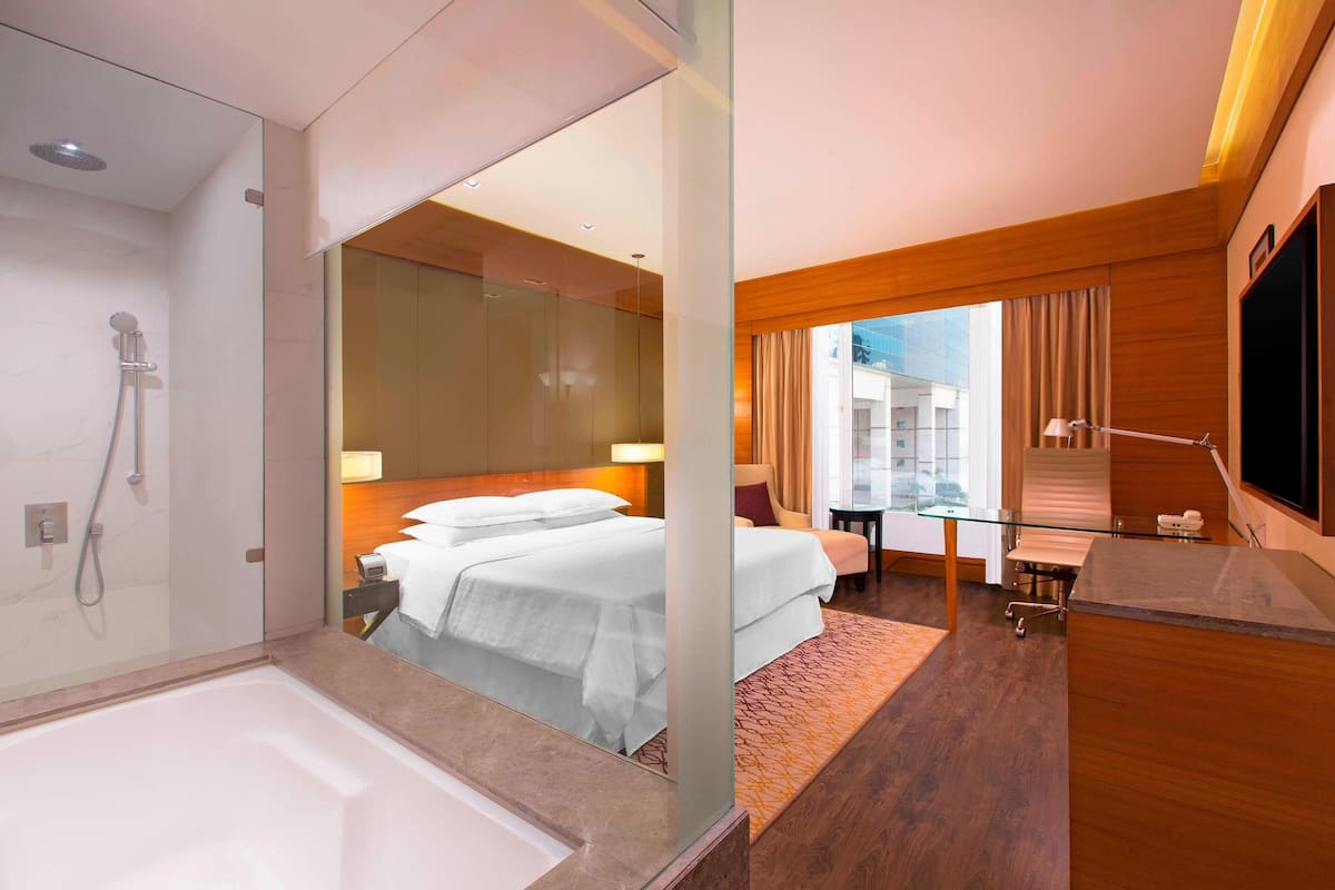 Ein modernes Hotelzimmer mit Duschkabine aus Glas, einem großen Bett mit weißer Bettwäsche, einem Schreibtisch mit Stuhl und einem an der Wand montierten Fernseher.