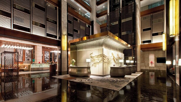 Een ruime lobby met een centraal receptiebalie, een zithoek met banken en een decoratieve wand met een groot kunstwerk.