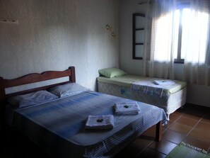 A bedroom with two beds, one larger and one smaller, both with striped bedding. There is a window with curtains, a wall-mounted shelf with decorative items, and a tiled floor. - Itaúna Hostel (Saquarema)