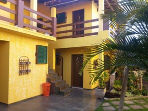 A two-story building with a yellow facade, a wooden balcony, and a small garden area with a palm tree.