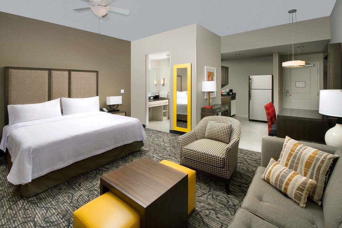 Een hotelkamer met een bed, een bank, een stoel, een klein tafeltje, een spiegel, een bureau en een koelkast.