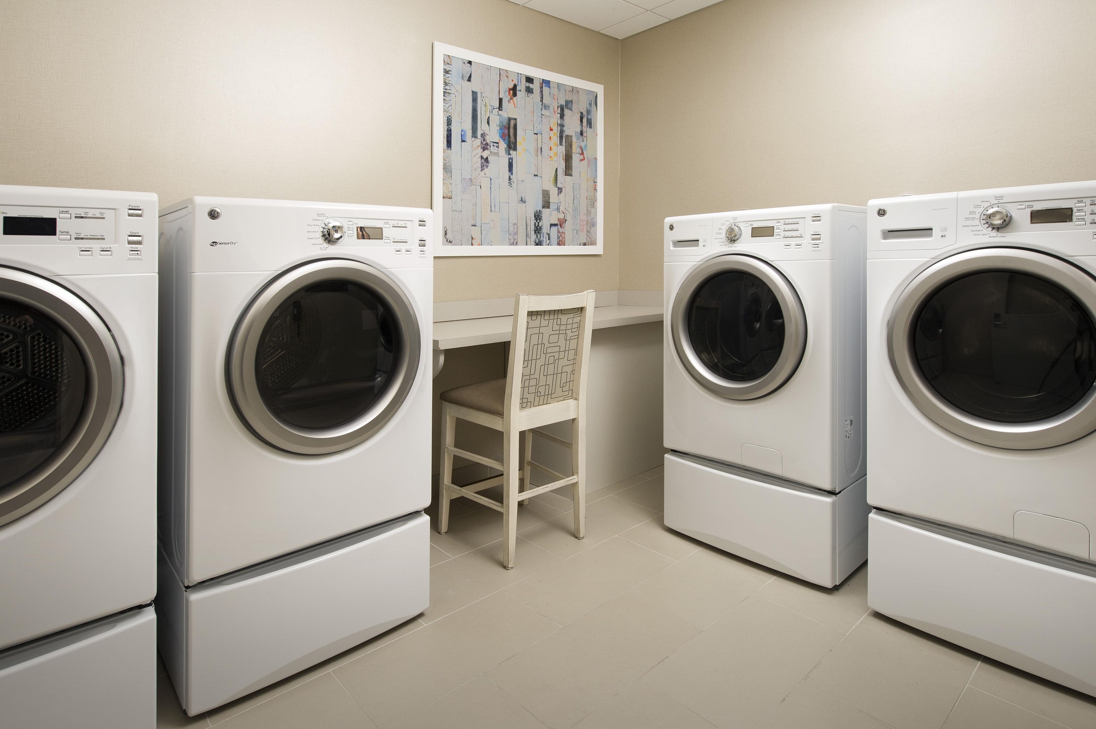A laundry room with three stacked front-loading washing machines, a chair, and a wall-mounted artwork.