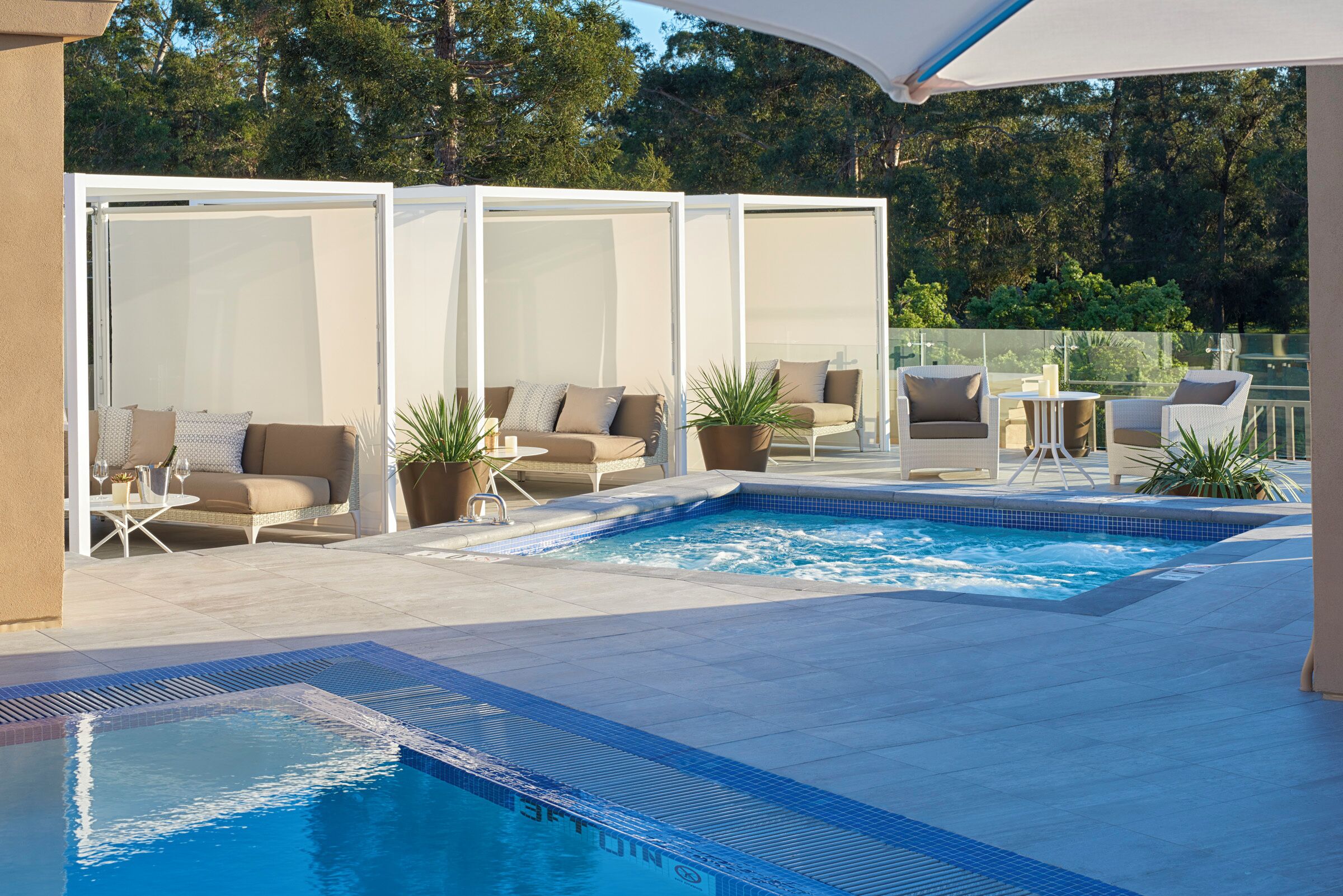 An outdoor pool area with lounge chairs and a small table, surrounded by a white fence and potted plants.