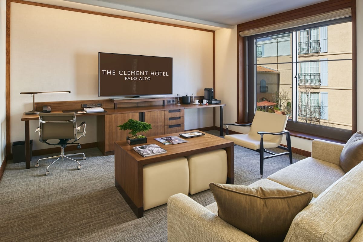 A hotel room with a TV, desk, office chair, sofa, and a window with a view of buildings.