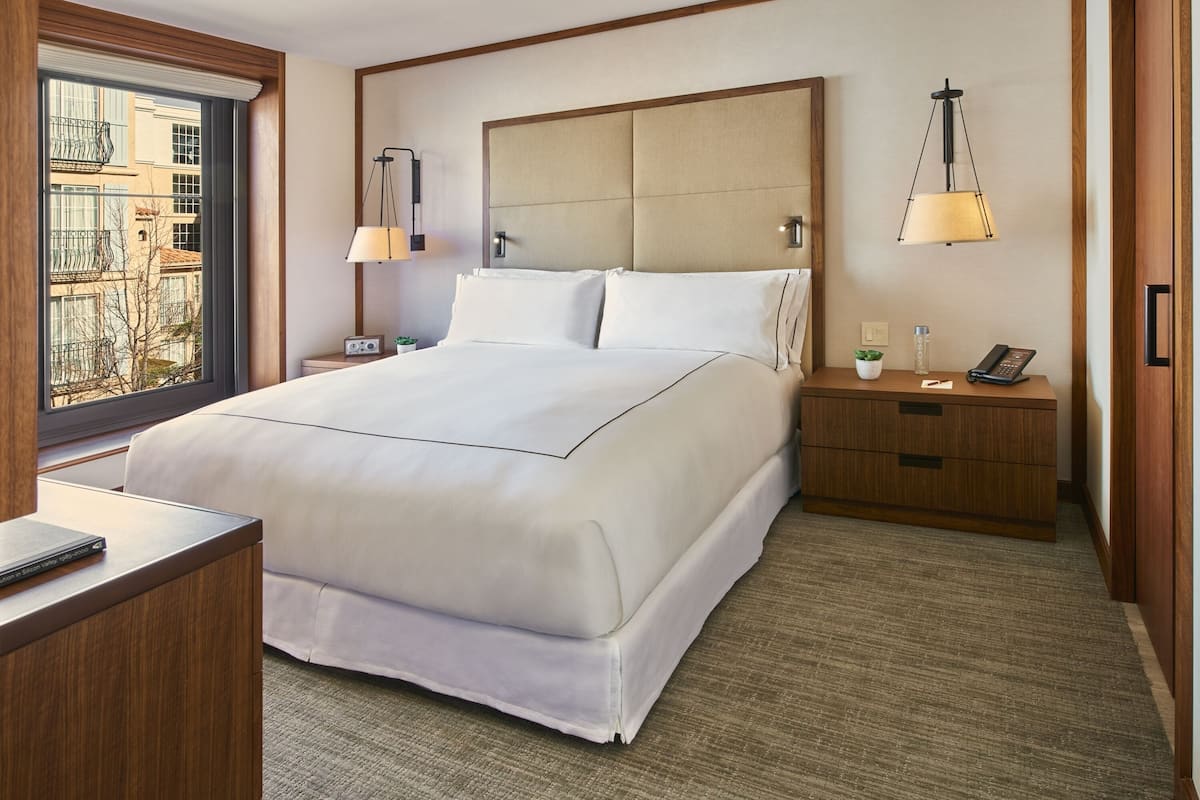 A hotel room with a large bed, bedside tables, a nightstand, a lamp, a phone, and a window with a view of buildings.