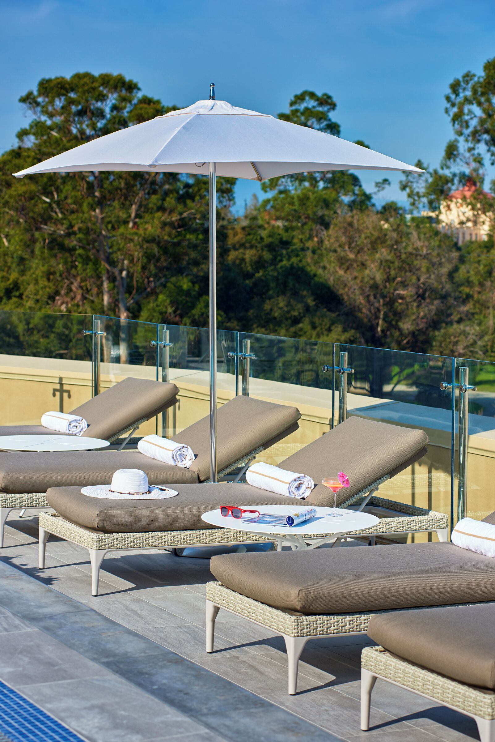 Un área de lounge en la azotea con sillones de sol, una sombrilla blanca y barandilla de cristal.