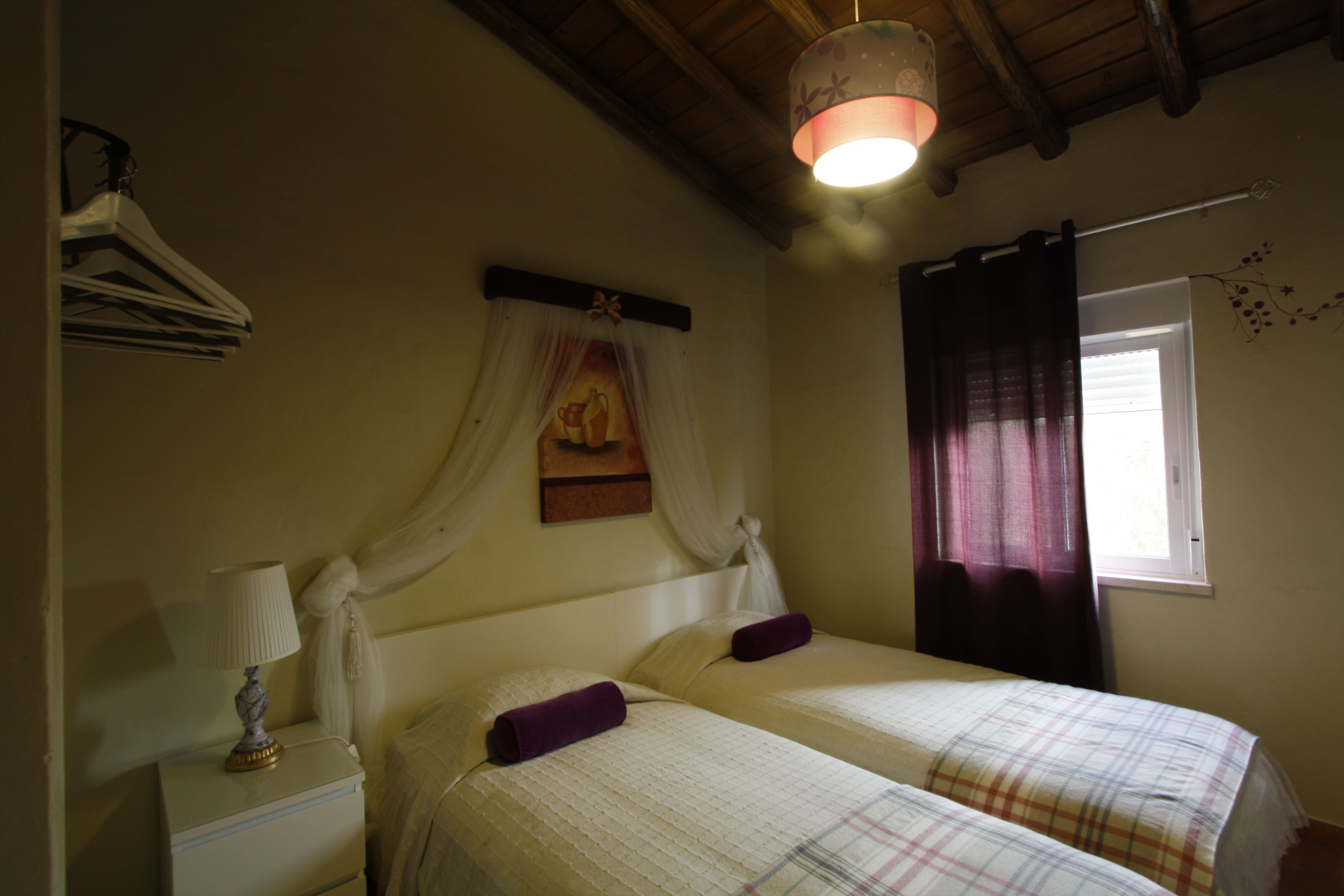 A bedroom with two beds, a nightstand with a lamp, a window with curtains, and a wall-mounted light fixture.