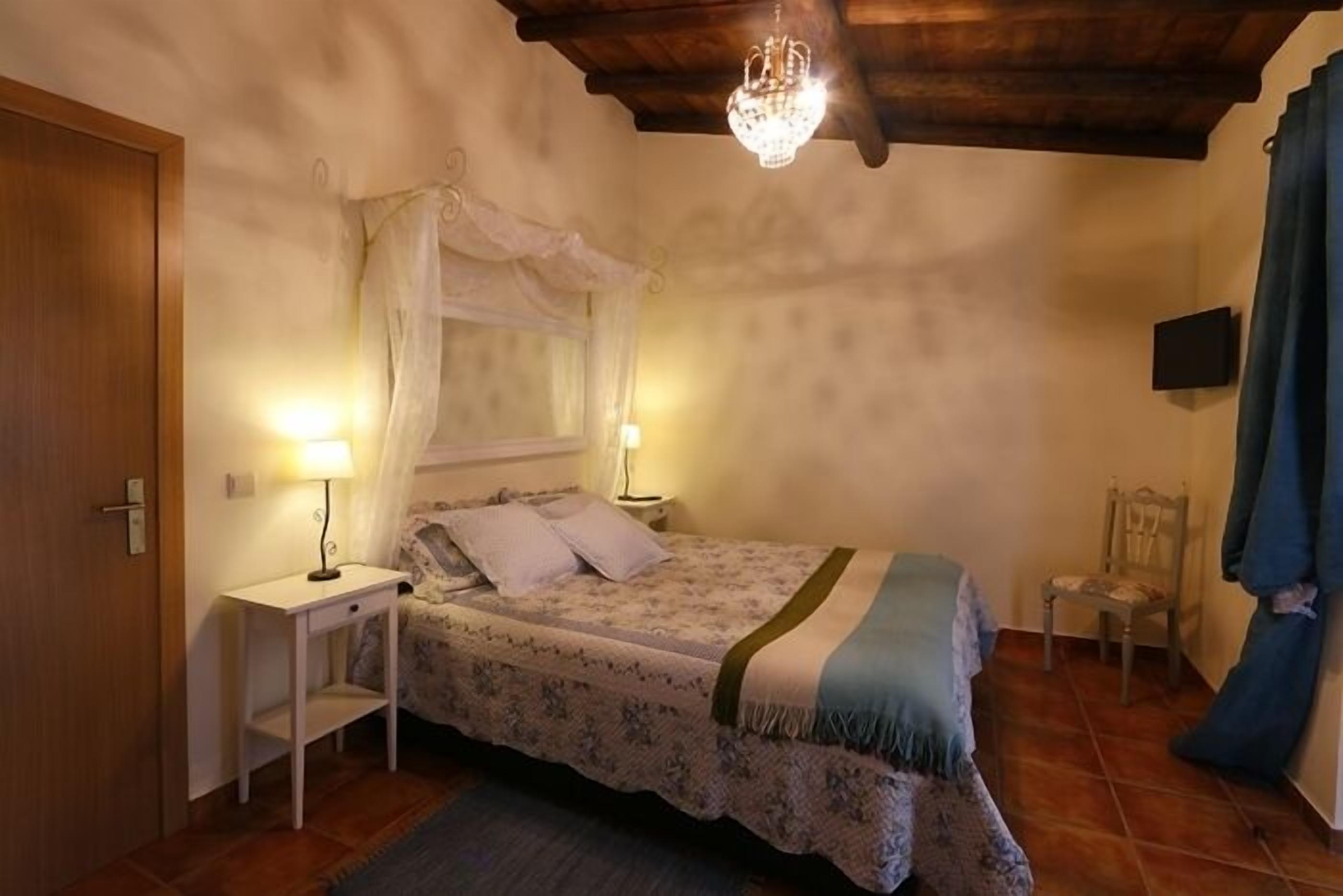 A bedroom with a bed, bedside tables, a chandelier, a window with a curtain, and a television on the wall.