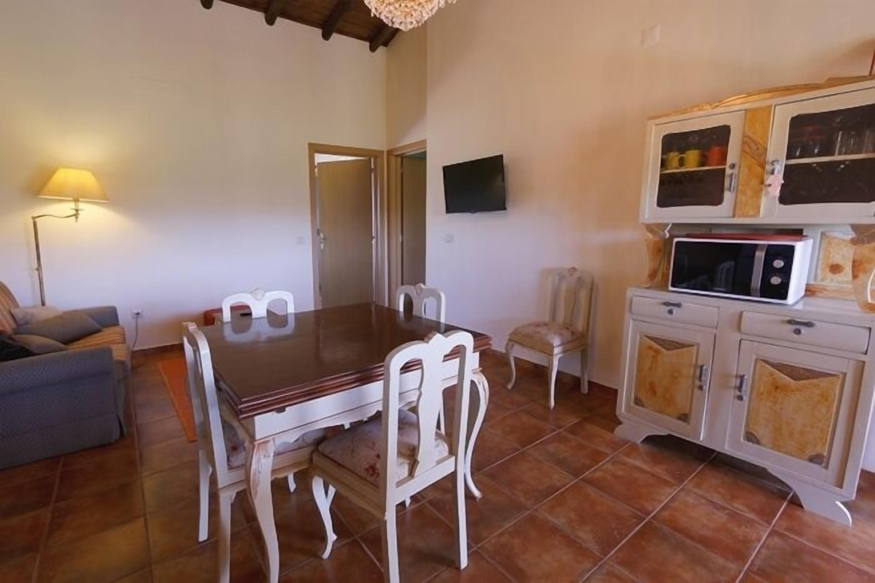 Uma área de refeições com mesa de madeira e cadeiras brancas, um sofá, uma televisão e um armário com portas de vidro.