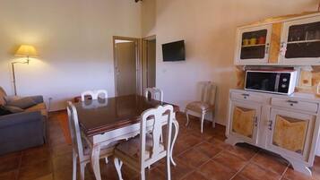 Uma área de refeições com mesa de madeira e cadeiras brancas, um sofá, uma televisão e um armário com portas de vidro.