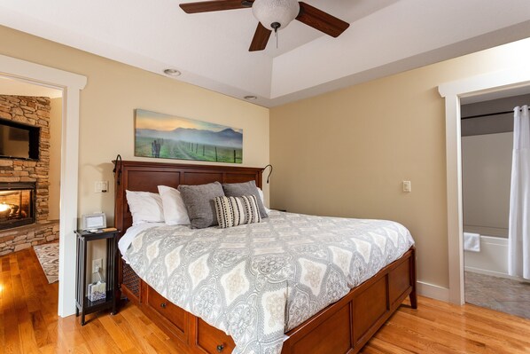 A bedroom with a wooden bed, a stone fireplace, a ceiling fan, and a painting on the wall. - Asheville Cottages (Asheville)