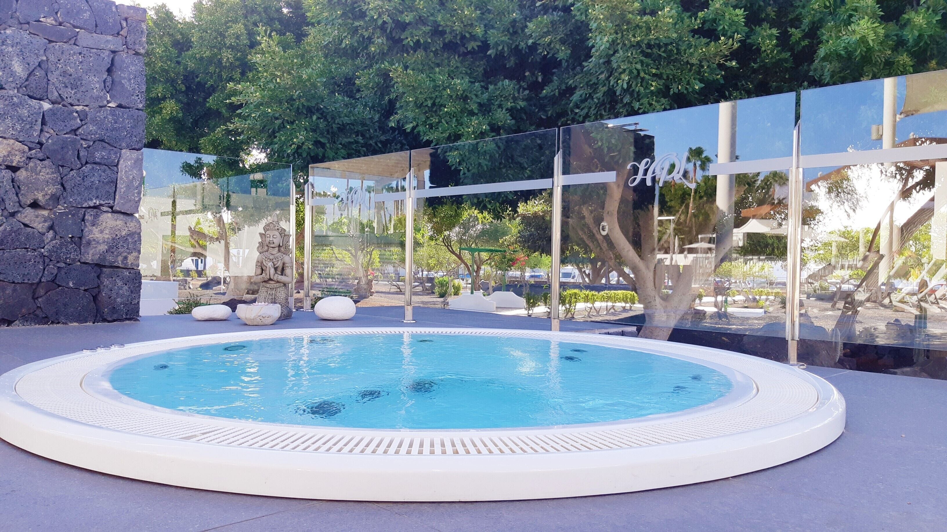 A hot tub with a stone wall and glass panels, surrounded by greenery and a pool.