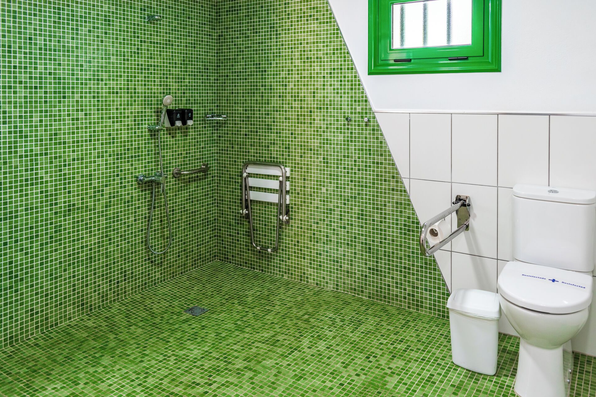 Une salle de bain avec des carreaux de mosaïque verts, des toilettes blanches, une poubelle blanche et une fenêtre à cadre vert.