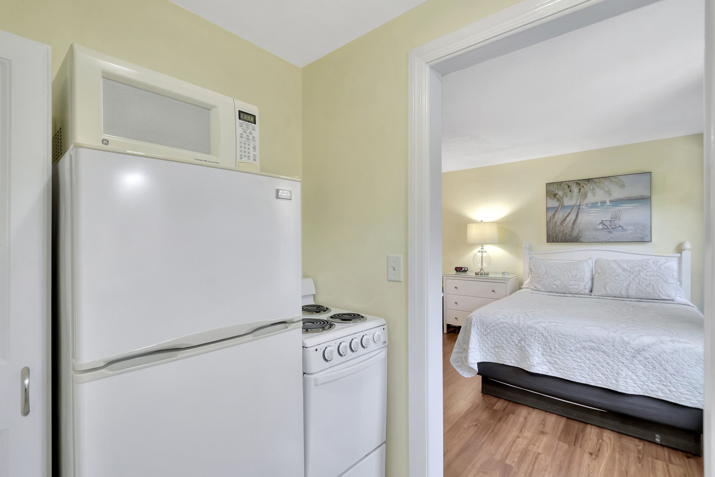 A compact kitchen with a refrigerator, stove, and microwave. Adjacent is a bedroom with a bed, nightstand, and a painting on the wall.