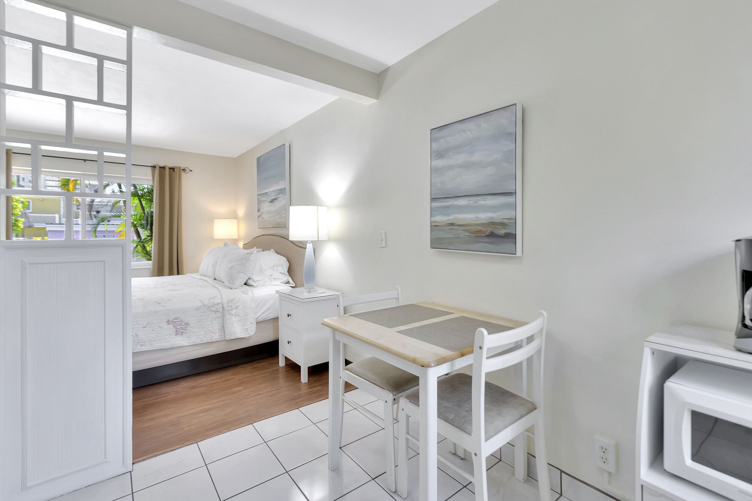 A hotel room with a bed, bedside table, lamp, and a small table with a chair. There are paintings on the walls and a view of the outdoors through a window.