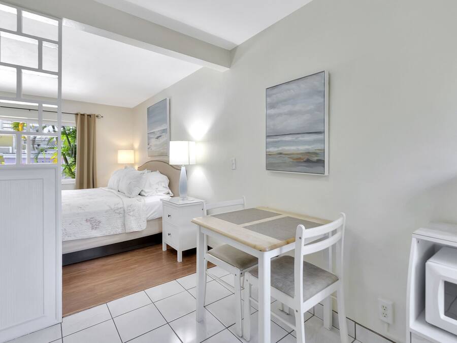 A hotel room with a bed, bedside table, lamp, and a small table with a chair. There are paintings on the walls and a view of the outdoors through a window.