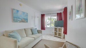 Ein Wohnzimmer mit einem beigen Sofa, einem Fernseher auf einem Schrank, einem Gemälde und einem Fenster mit roten Vorhängen.