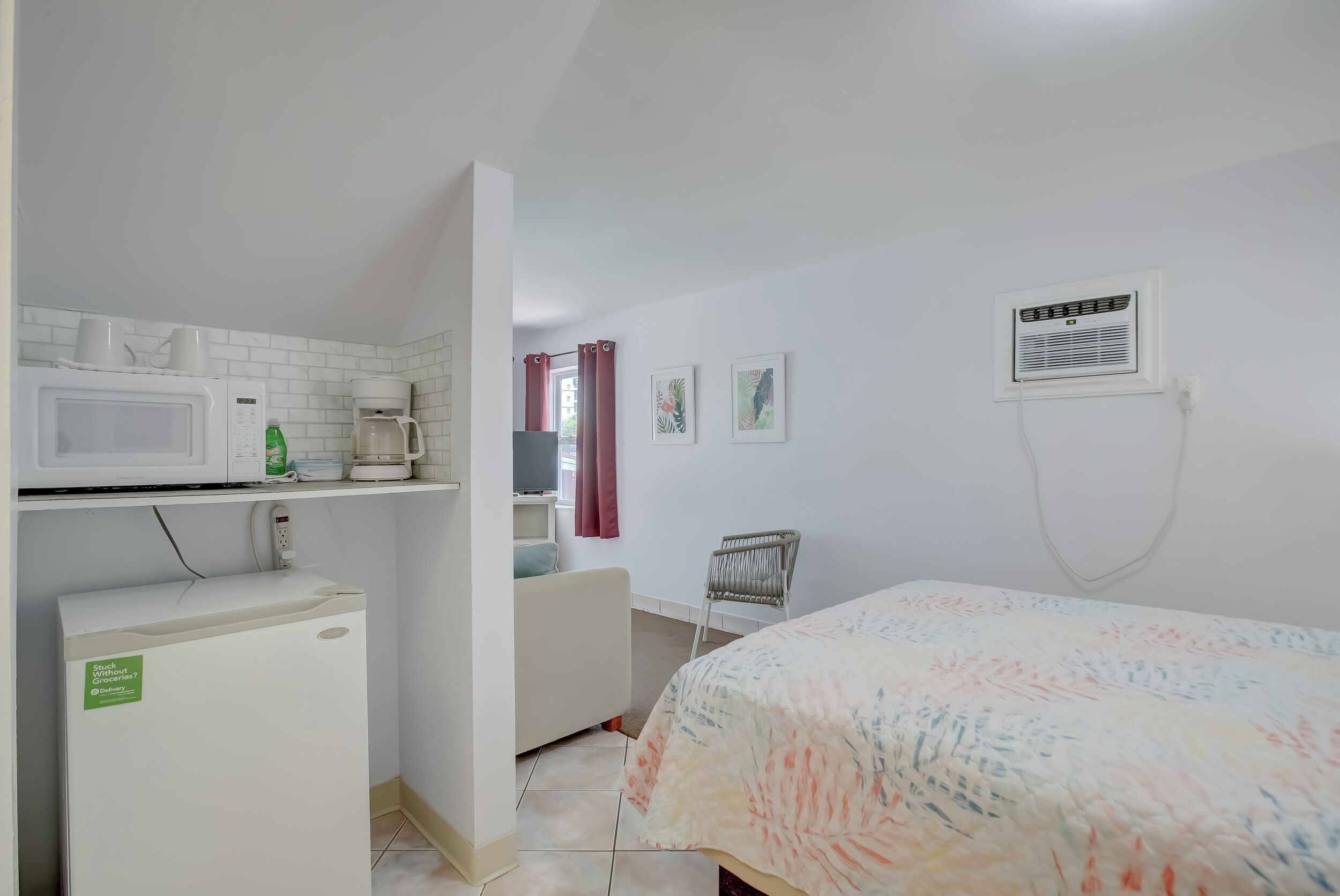 A compact room with a bed, a small kitchenette with a microwave and refrigerator, a chair, and a window with curtains.