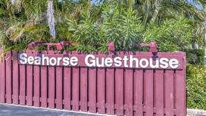 Ein Holzschild mit dem Schriftzug 'Seahorse Guesthouse' vor einer Kulisse aus Palmen.