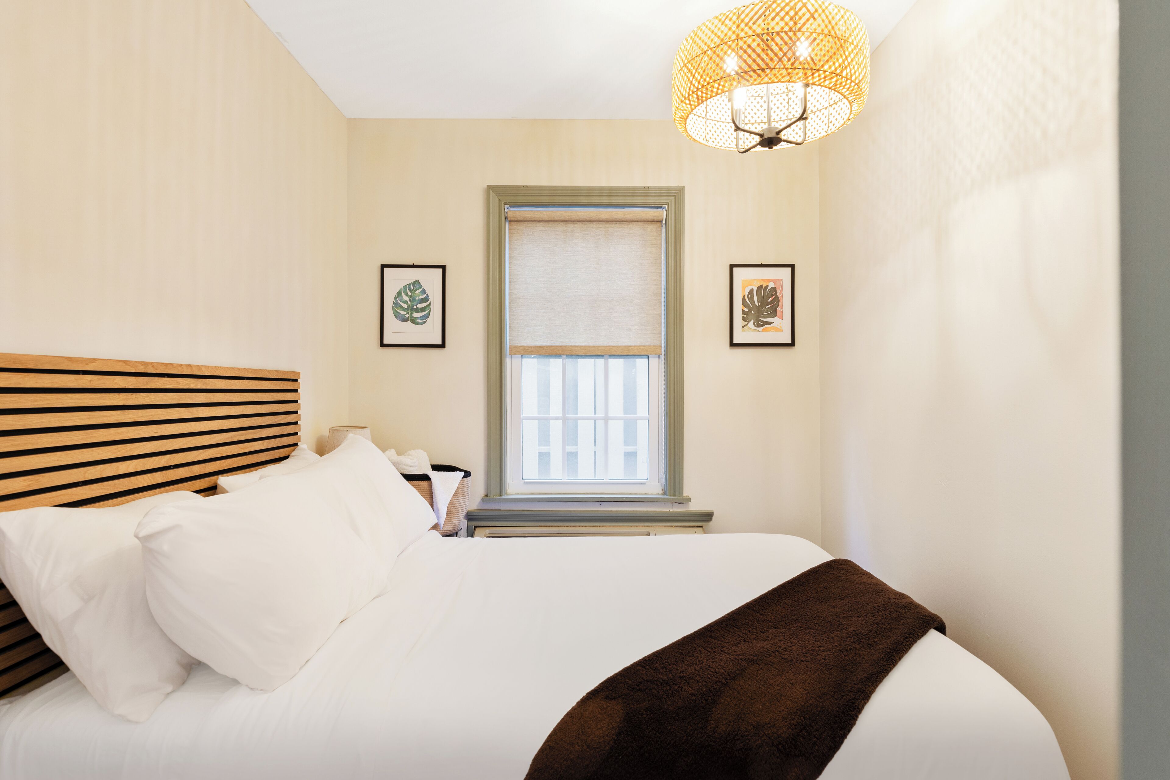 Un dormitorio con cama, ventana con persianas, lámpara de pared y cuadros enmarcados.