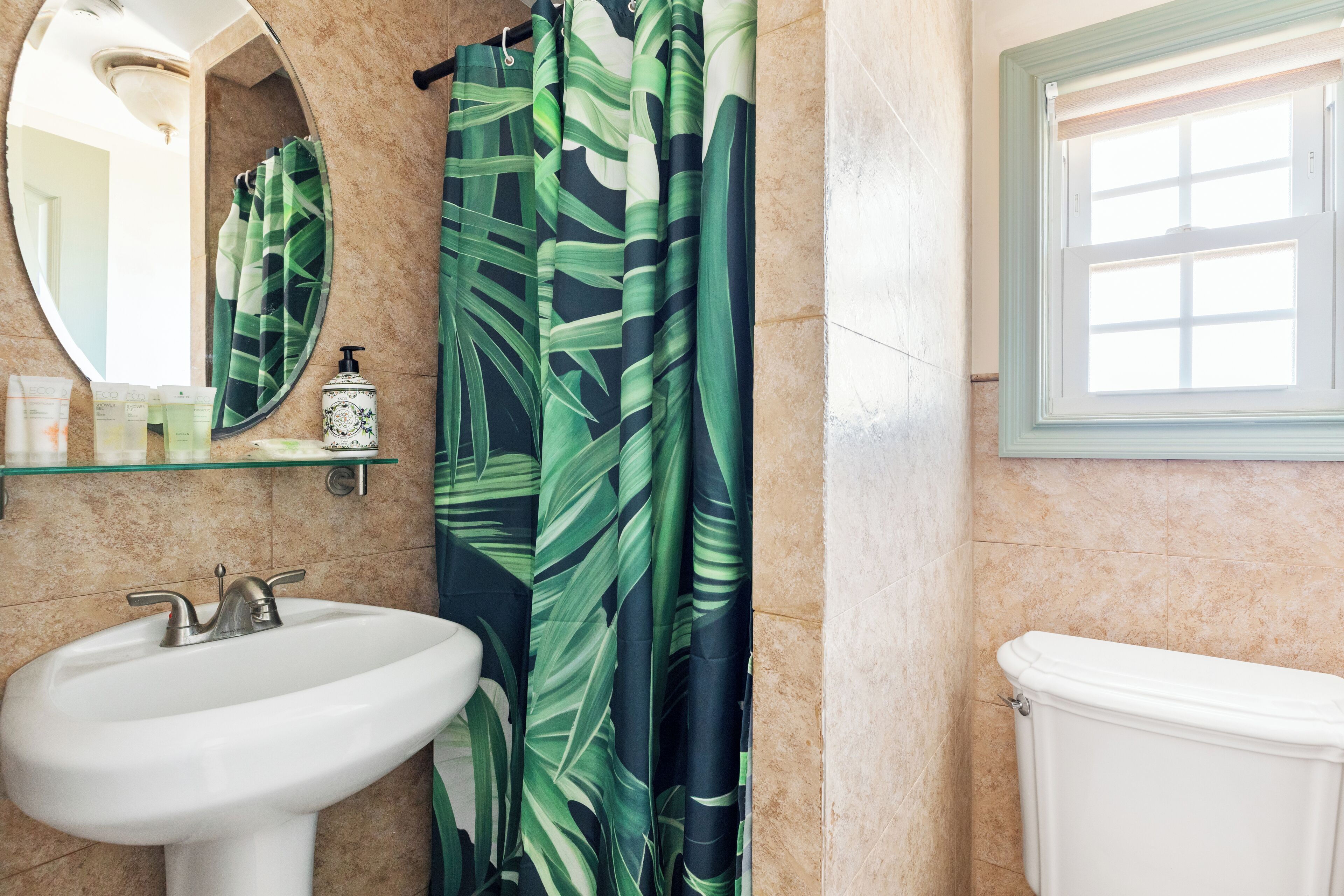 Un baño con lavamanos blanco, un espejo, un inodoro y una cortina de ducha estampada con hojas.